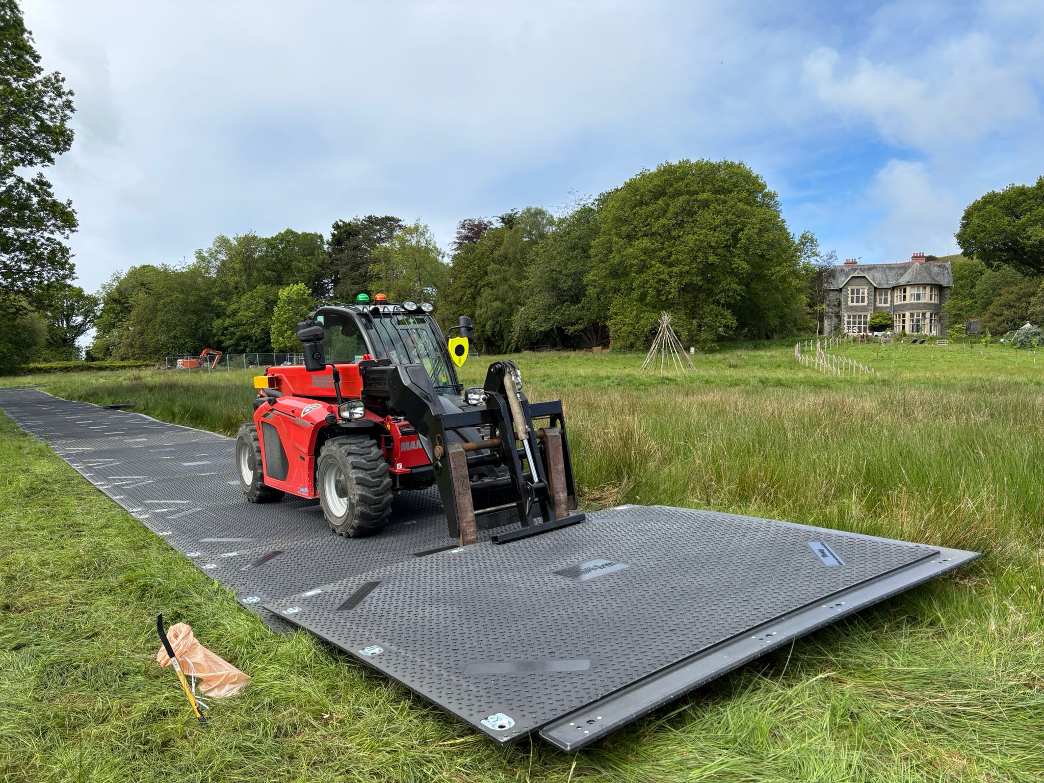 Red telescopic handler placing large metal trackway panels on grassy field with trees and house in background.